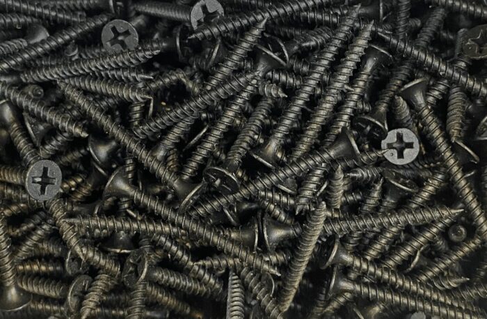 A top-down, close-up view of a large pile of black gypsum screws with Phillips heads, scattered randomly.
