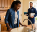 A photograph of a frustrated Kenyan woman in her kitchen, looking at a dripping faucet with low water pressure, while a calm plumber stands behind her with a toolbox and tablet.